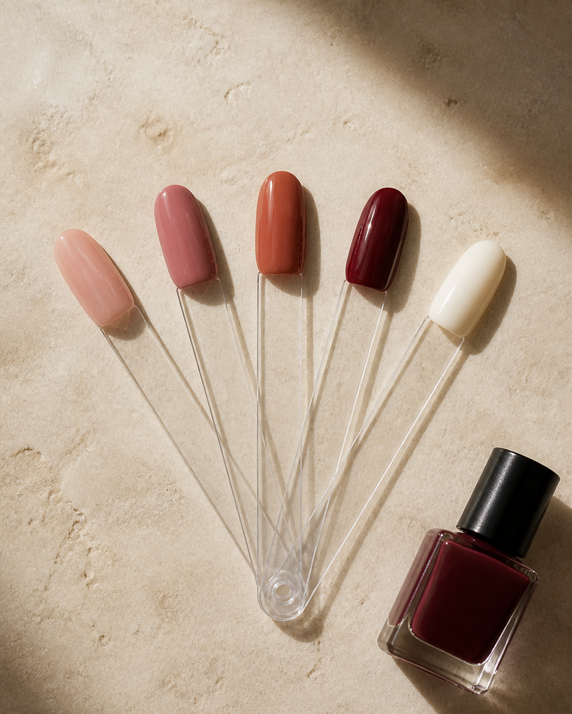 Nail color samples in soft pink, terracotta, burgundy, and cream arranged on a warm stone surface